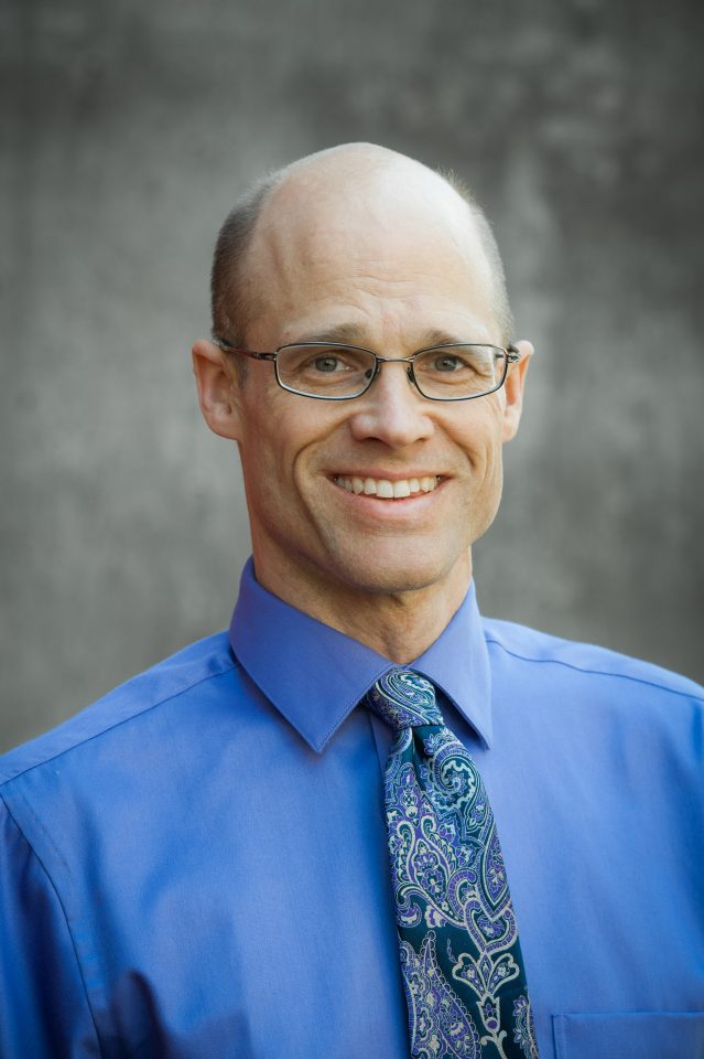 A headshot of Dr. Mortenson wearing a blue shirt and tie